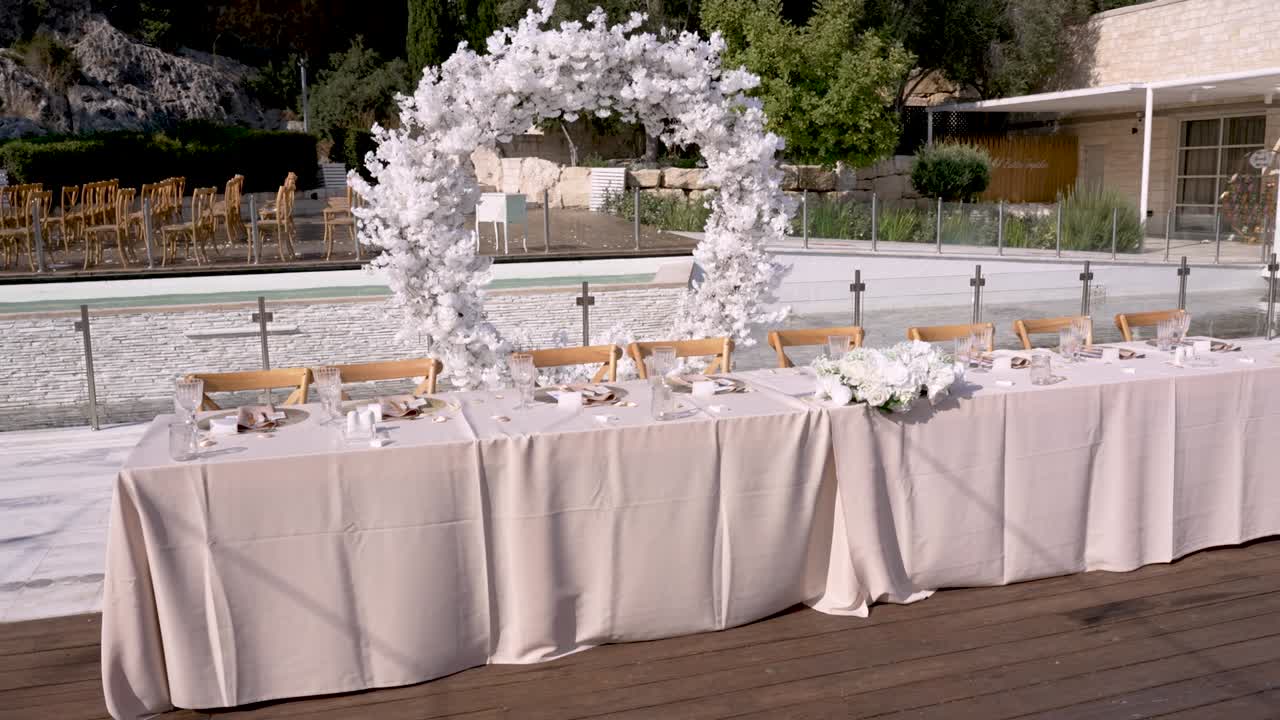 Un bellissimo ricevimento nuziale all'aperto è allestito con una splendida tavola, decorazioni floreali bianche e uno sfondo sereno. La scena include una piscina scintillante e la natura.