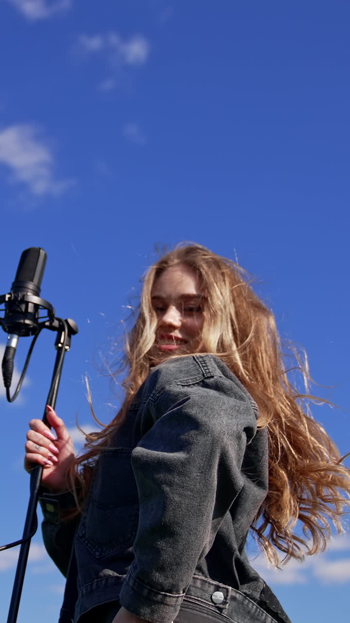 Pop singer woman on sky background. Beautiful girl in jeans clothing dancing and singing into the microphone on field in sunny day. Vertical video