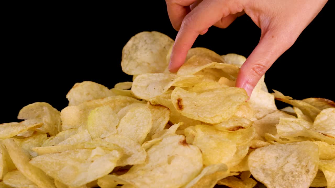 Human hand grabs potato chips from pile, studio lighting, close-up, black background, static camera