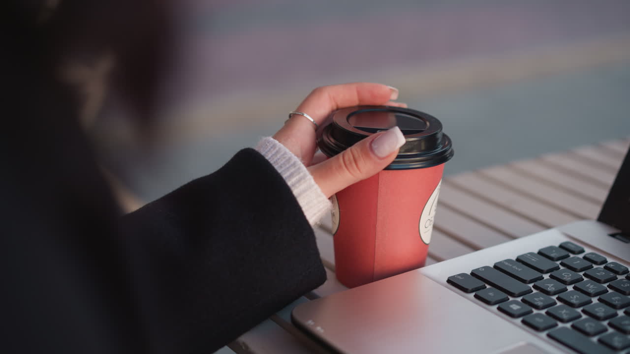 primer plano de la mano de la dama girando suavemente la taza de café con los dedos cuidados, la computadora portátil borrosa en el fondo creando un ambiente de trabajo sofisticado
