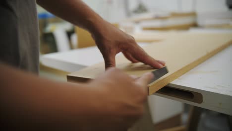 The image shows a person using a hand file to smooth the edge of a wooden board in a workshop. One hand steadies the board while the other works with the file.