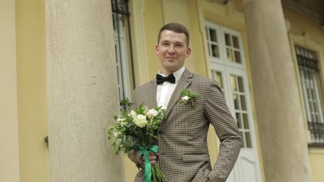 novio, joven quedarse cerca de la casa con columnas con ramo de bodas. él esperando a su amada novia