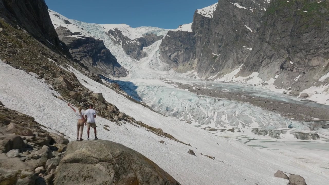 Un drone cinematografico riprende una coppia in piedi in una vasta valle che si affaccia sul ghiacciaio Austerdalsbreen, Norvegia, rivelando drammatiche texture di ghiaccio glaciale e motivi naturali sotto un cielo blu