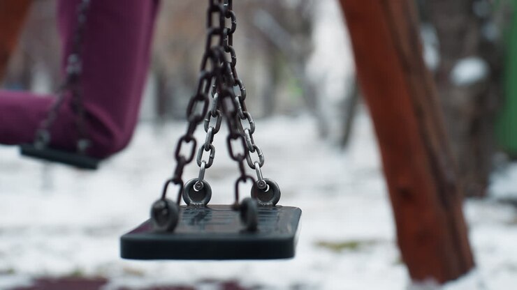 Vista parziale di una persona con pantaloni bordeaux e stivali neri che si dondola delicatamente su un'altalena, con l'altalena in quiete, neve tutt'intorno e uno sfondo sfocato di alberi e strutture.