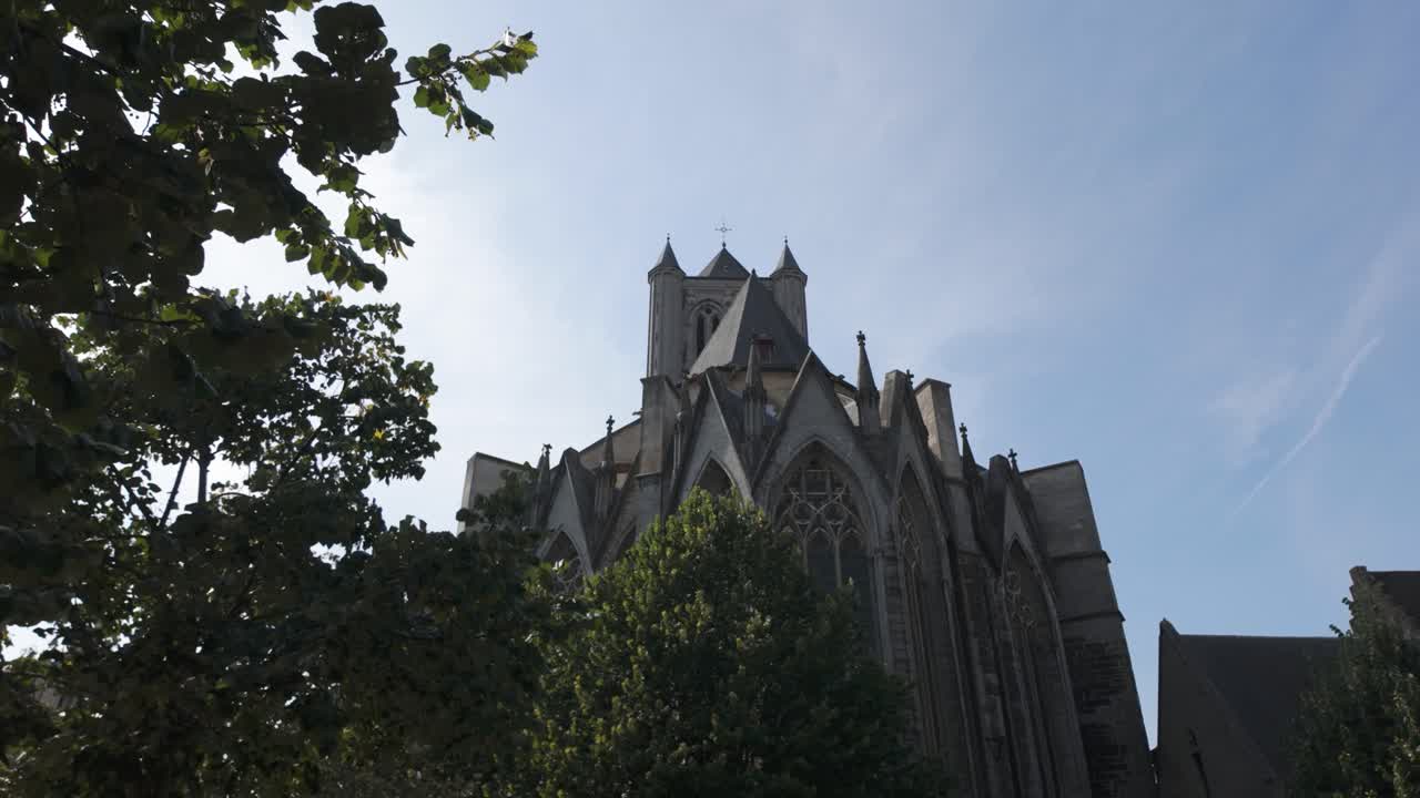 벨기에 겐트에 있는 성 니콜라스 성당의 풍경, 은 푸른 하늘 아래 초록색으로 둘러싸여 있습니다. 고 양식의 건축물은 조용한 나무와 조화롭게 조화를 이루고 있습니다.