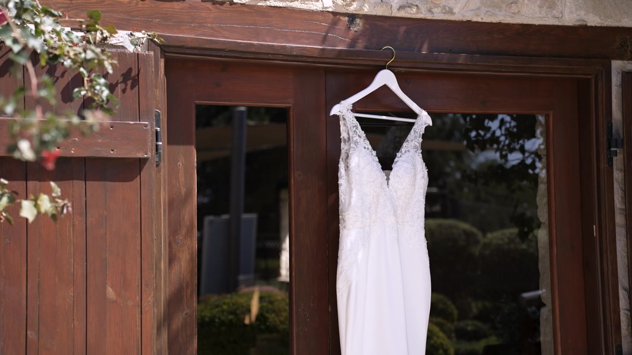 A wedding dress is displayed on a hanger outside a venue. The setting shows a rustic building with wooden trim and greenery. Sunlight shines on the dress, highlighting its details.