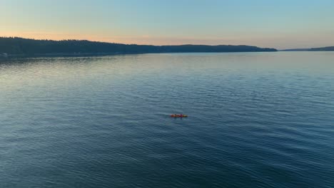 Vista-Aérea-De-Drones-Dando-Vueltas-A-La-Izquierda-De-Dos-Personas-En-Kayak-De-Mar-Remando-En-Una-Bahía-Cerca-De-Seattle,-Washington-Al-Amanecer,-Revelando-La-Ciudad-De-La-Comunidad-De-Southworth