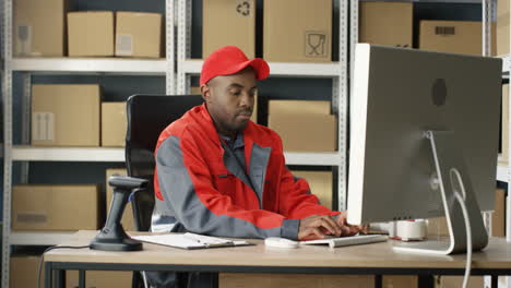 Joven-Cartero-Guapo-En-Uniforme-Y-Gorra-Sentado-En-La-Computadora-Y-Llenando-La-Factura-Del-Paquete