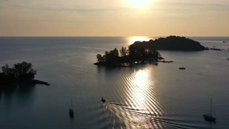 Cinematic-aerial-sunset-landscape-with-glowing-sun-setting-on-the-horizon,-boat-sailing-on-calm-sea-waters-with-golden-sunlight-reflection,-tropical-paradise-at-Langkawi,-Kedah,-Malaysia