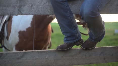 close shot of the boots of a woman sitting on the wooden fence of a ranch with a horse behind in slow motion