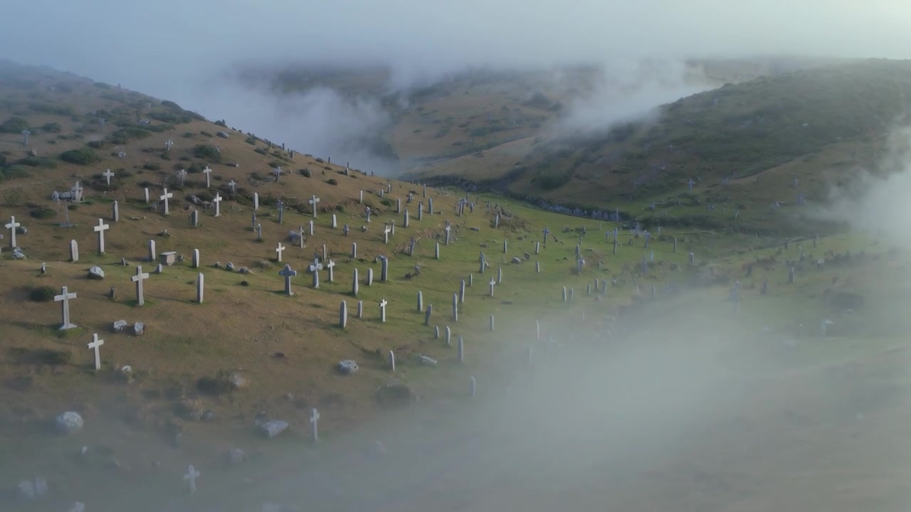 Telecamera che si allontana mentre la nebbia si dirada, filmando un cimitero in collina con croci per mostrare le tombe.