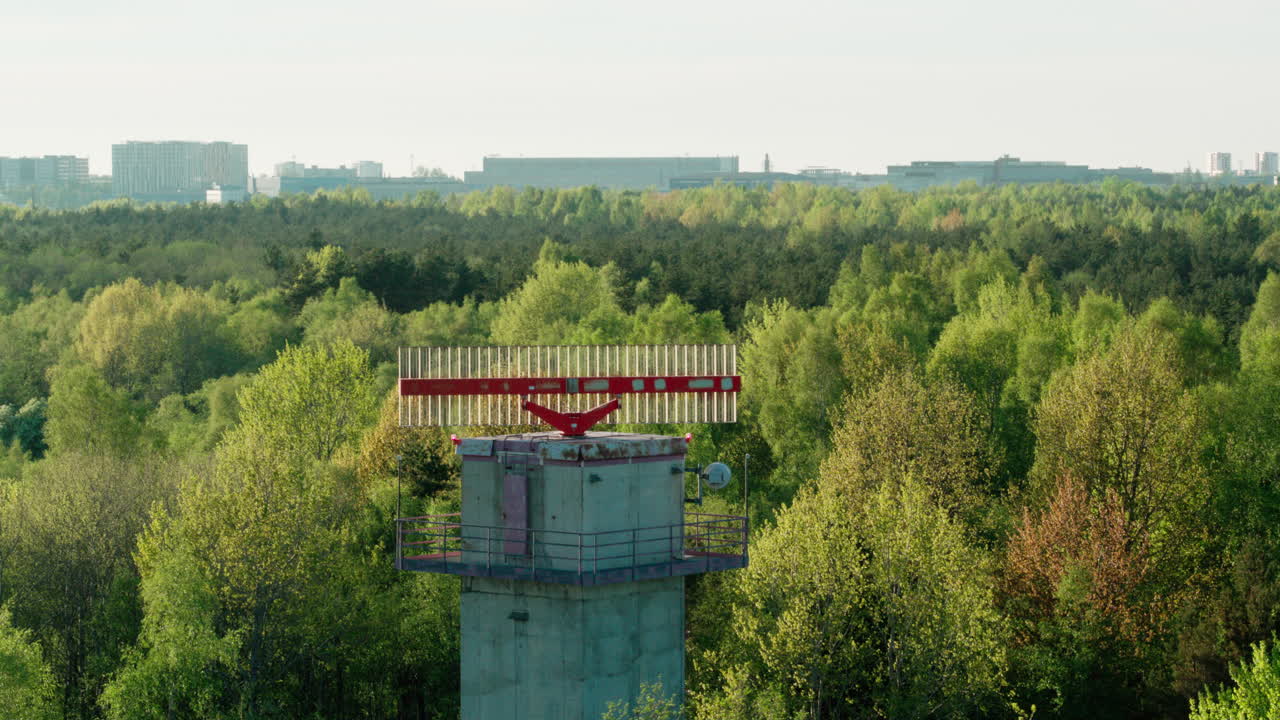 Old and worn secondary surveillance radar for air traffic control rotating in the middle of forest with city visible in background. Wider shot.