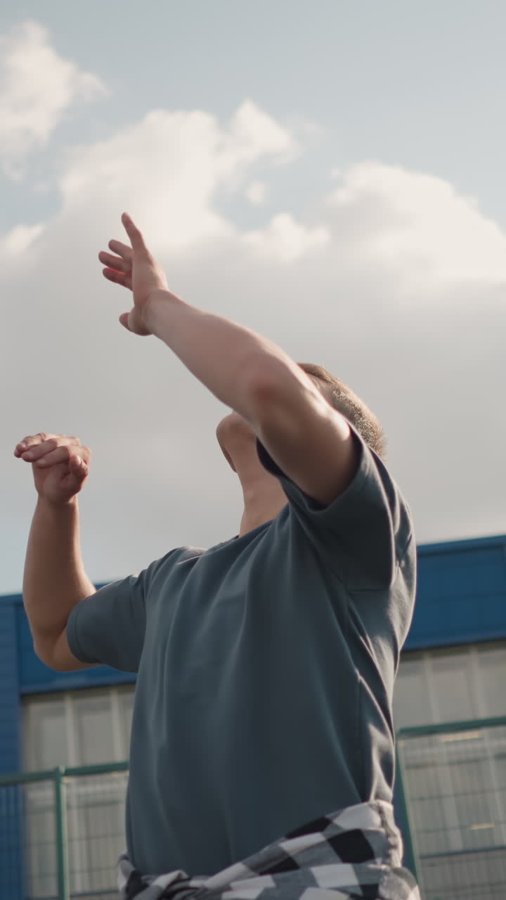 joven atleta girando la pelota de voleibol en la mano antes de lanzarla y servirla cerca de la cancha de baloncesto, con edificio azul y vegetación en el fondo