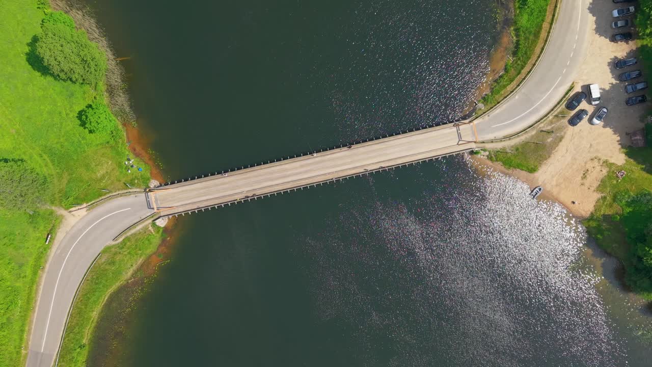 Aerial top down view of a road bridge crossing a river with parked cars, green fields, and forest on a sunny summer day