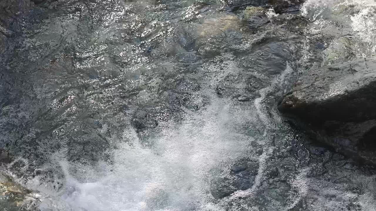 Fast-moving clear water rushes over dark river rocks in natural daylight, captured with a steady overhead camera angle