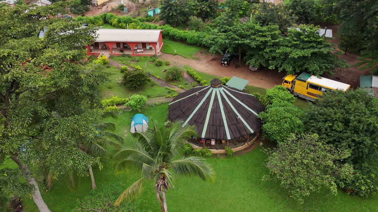 A bird's-eye drone shot over a lush green eco-resort in the Volta Region of Ghana. Features traditional circular buildings, camping tents, and a travel bus nestled in nature
