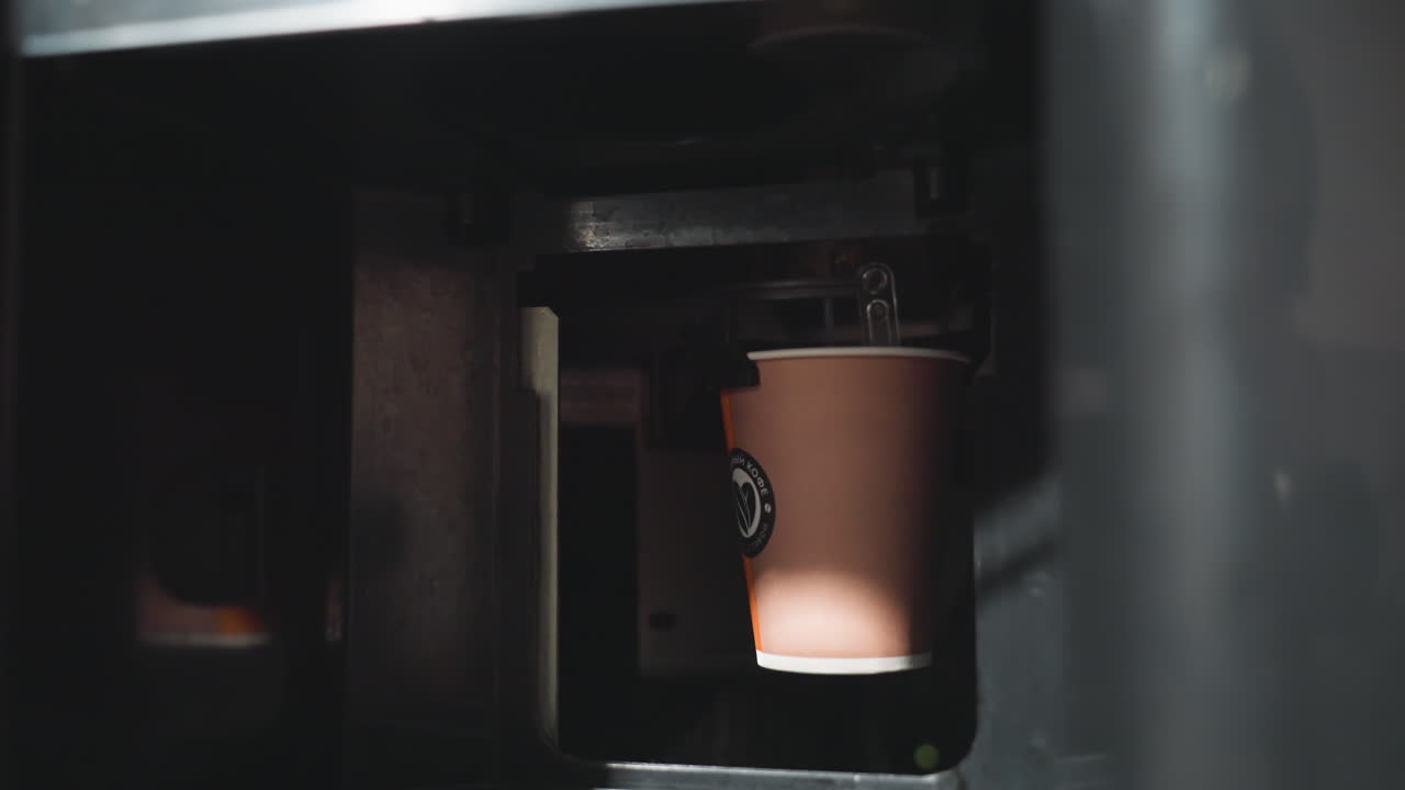 Dispensador de vasos de papel en una máquina expendedora, mostrando el café que se sirve dentro de una máquina de bebidas automática, capturando el vaso en movimiento mientras cae sobre una plataforma de metal en un entorno de oficina moderno