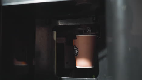 paper cup dispensing from vending machine, showing coffee being served inside automatic beverage machine, capturing cup in motion as it drops down on metal platform in modern office setting