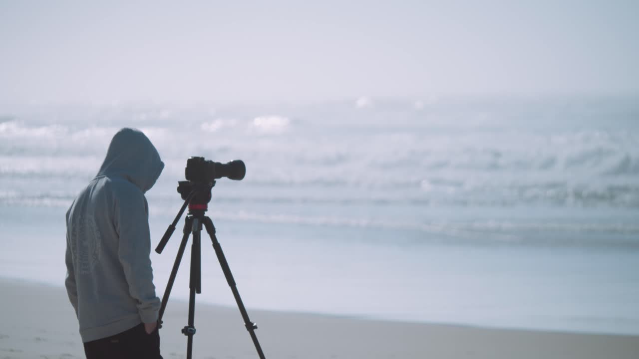 A cameraman filming or photographing the ocean from the beach in a hoodie