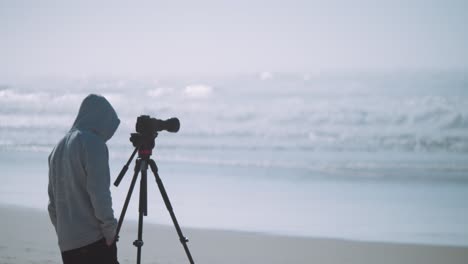 A cameraman filming or photographing the ocean from the beach in a hoodie
