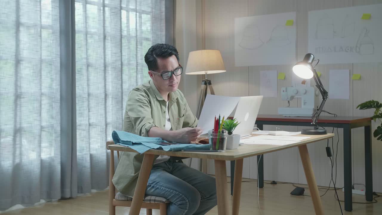 사무실에서 노트북으로 작업하는 동안 레이아웃 본드를 바라보는 직물로 된 아시아인 디자이너