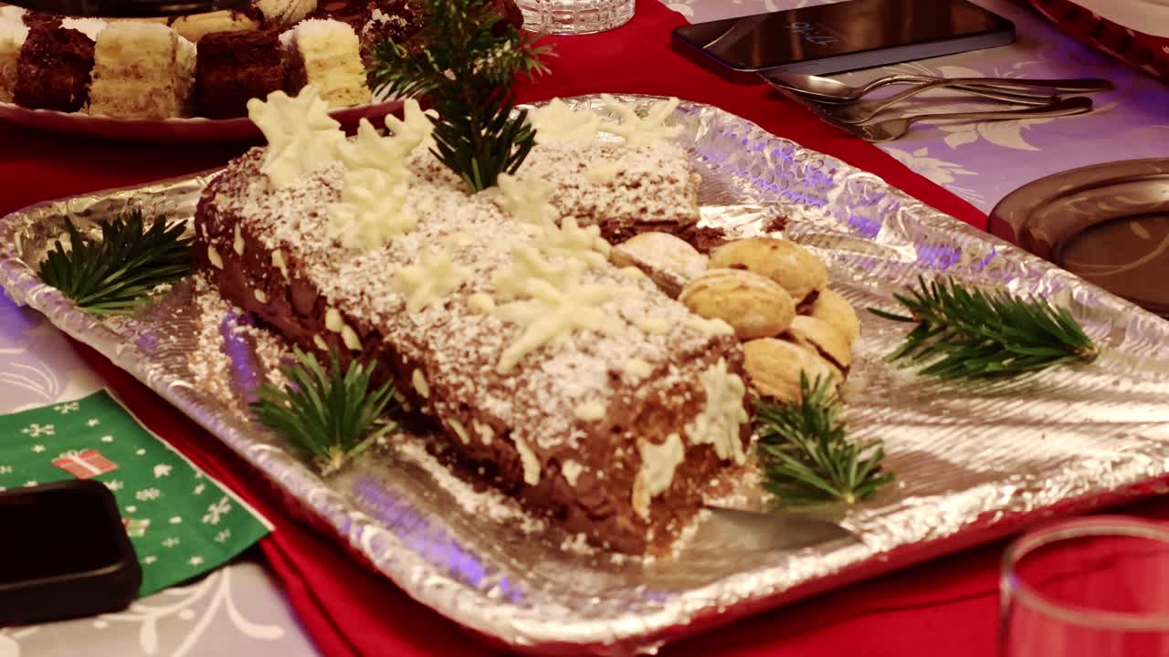 Nahaufnahme eines Weihnachtsstollenkuchens, der auf einem festlich gedeckten Tisch angeschnitten wird, dekoriert mit Puderzucker, Cremedetails, Walnusskeksen und immergrünen Zweigen