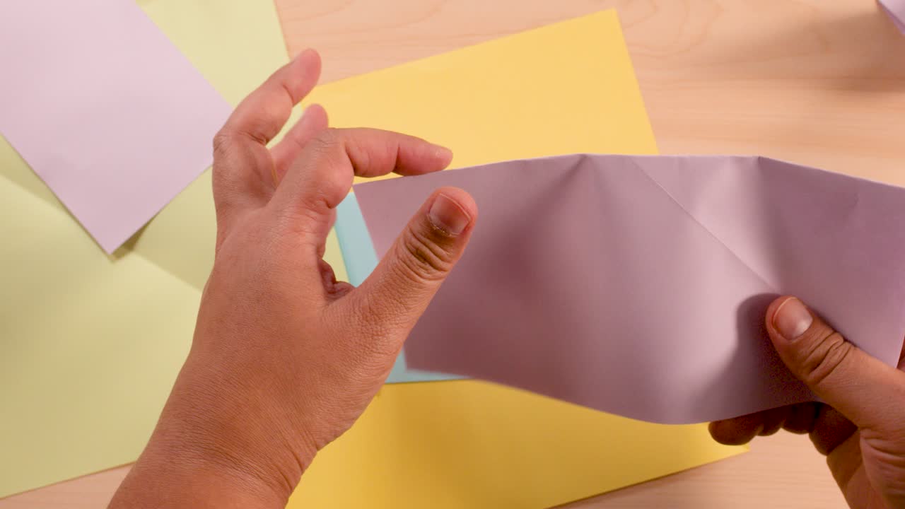 Hands methodically fold white paper on wooden desk under soft lighting, preparing for origami craft