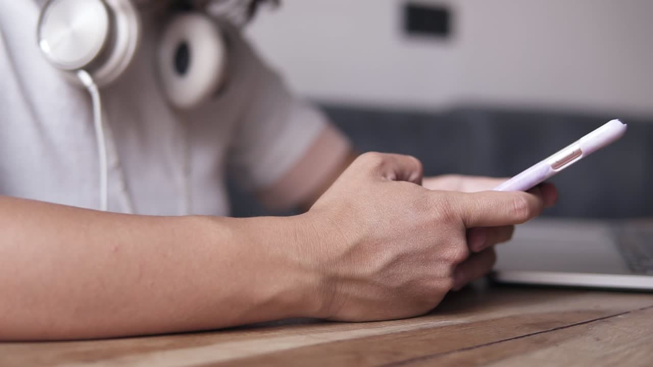 extremadamente cerca de una joven con camisa blanca sentada junto a la mesa con auriculares en el cuello. teléfono móvil en las manos, desplazándose por las páginas. sin rostro