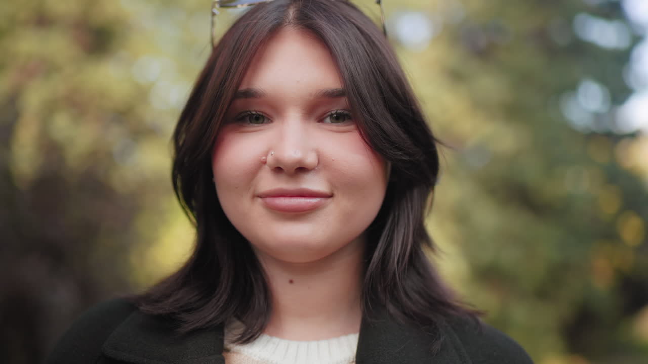 Крупный план: портрет красивой девушки с теплой улыбкой, солнечные очки на волосах, мягкий эффект боке на фоне листвы, уютное пальто и полосатый свитер, естественное освещение, непринужденное настроение на открытом воздухе, дружелюбное выражение лица