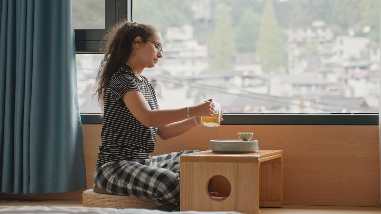 Rituale del tè alla finestra dell'hotel: donna seduta accanto alla finestra che sorseggia il tè a un tavolino basso, vista sulla città attraverso il vetro, top fantasia e pantaloni a quadri, tenda morbida, umore calmo e riflessivo, viaggiatrice solitaria che si gode un soggiorno lento.