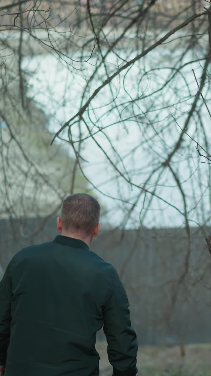 un hombre con una chaqueta verde está de pie en un bosque, tocando la corteza de un árbol con una mano mientras sostiene un par de auriculares en la otra