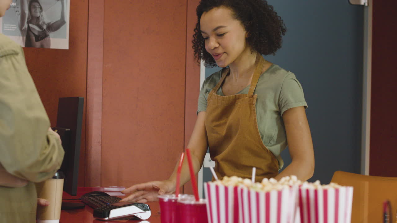 mujer sonriente vendiendo entradas de cine en el cine y el cliente pagando con una tarjeta sin contacto