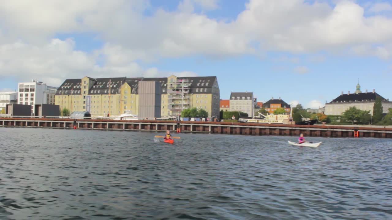 Floating on the water from right to left. Some people with kayaks paddling on the waterfront of Copenhagen, Denmark. Sunny day with cloudy sky.