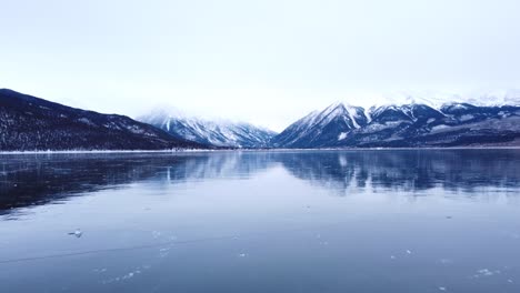 Flying-low-over-a-frozen-lake-and-then-climbing-to-a-higher-altitude-with-snow-capped-mountains-in-the-background,-aerial