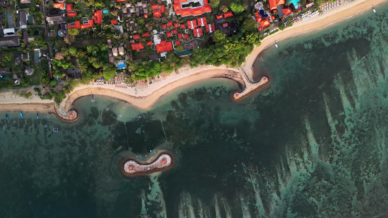 Aerial drone footage of Sanur Bali coastline with sandy shore, coral reef barriers, turquoise water, palm trees, coastal village, lush greenery, sunrise light, and peaceful tropical seascape