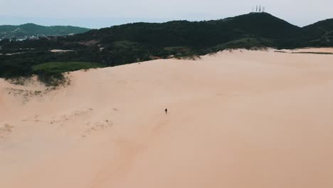 Sandboarder-walking-alone-on-big-sand-dunes-near-the-tropical-beach-of-Garopaba,-Santa-Catarina,-Brazil