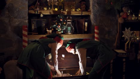 curious elves in small decorated room opening the magic box. two elves in fairy suits taking present from the chest in a dark christmas room.