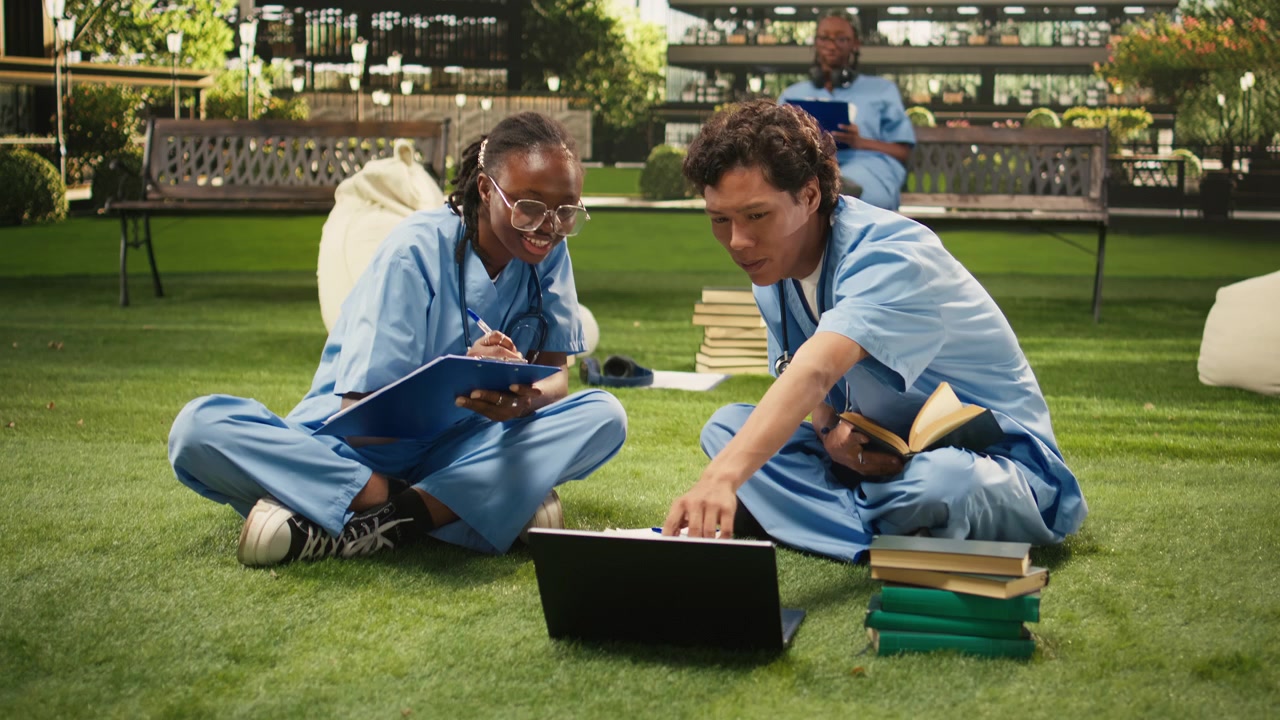 Medical students in blue scrubs discuss coursework beside laptop