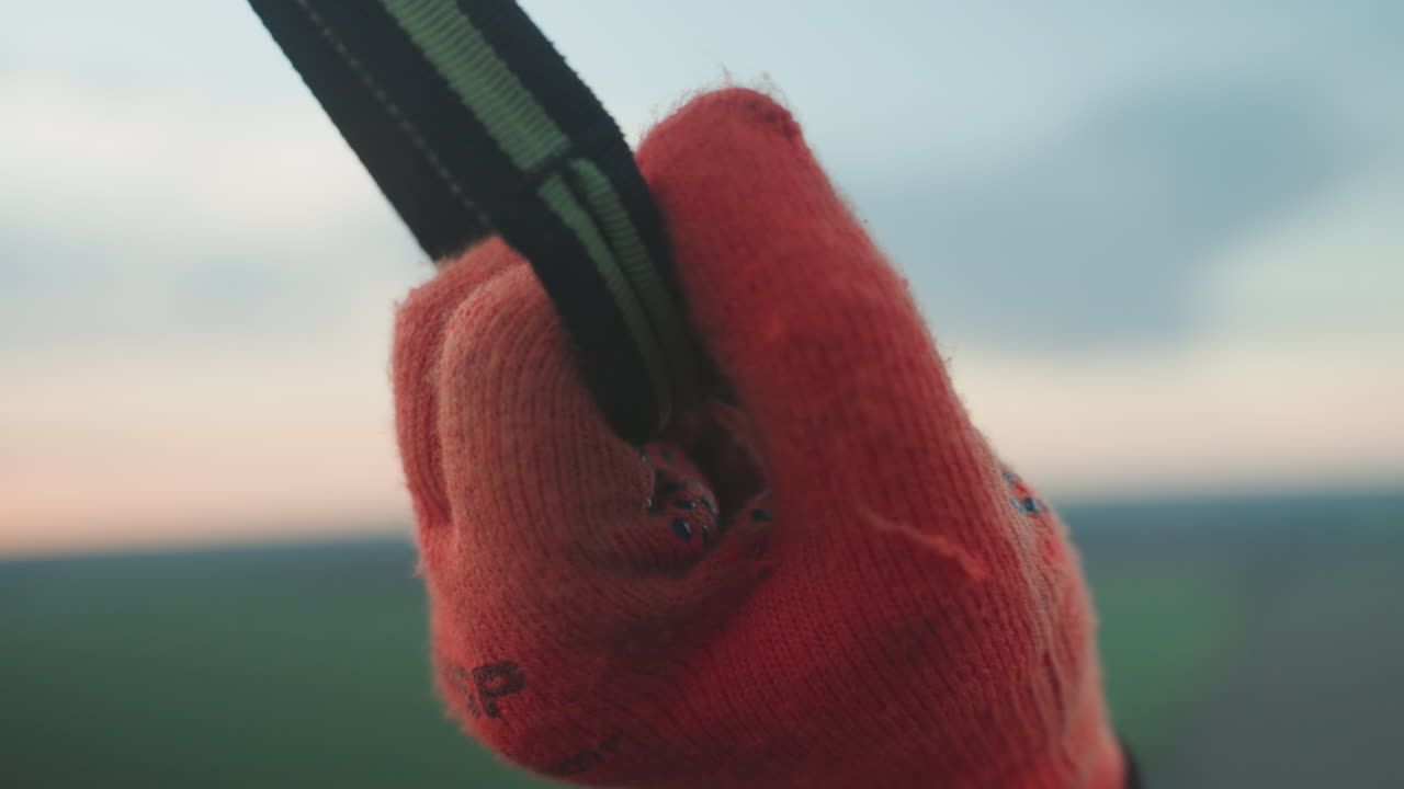 person in red glove grips black nylon strap of paramotor frame against blurry grassy field and pastel twilight sky with distant treeline while checking flight gear during prelaunch preparation