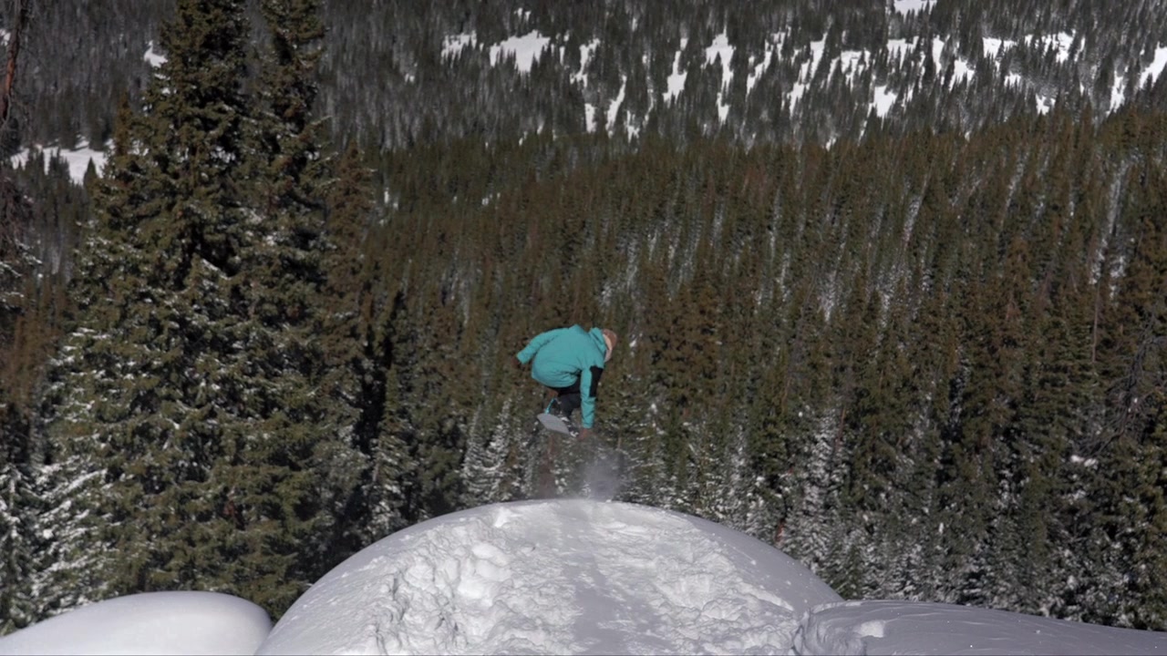 Snowboarder professionista di backcountry che esegue un salto da una scogliera a cuscino con trick aerei Indy grab, scendendo un terreno ripido. Inverno a Vail Pass, Montagne Rocciose del Colorado, con neve fresca e profonda, cielo blu e mattina soleggiata.