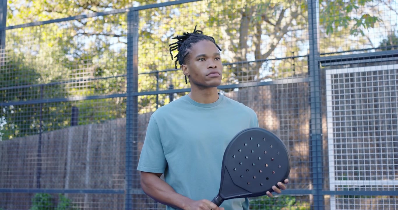 Adult African American man in teal tee examining black paddle on fenced court, preparing to play