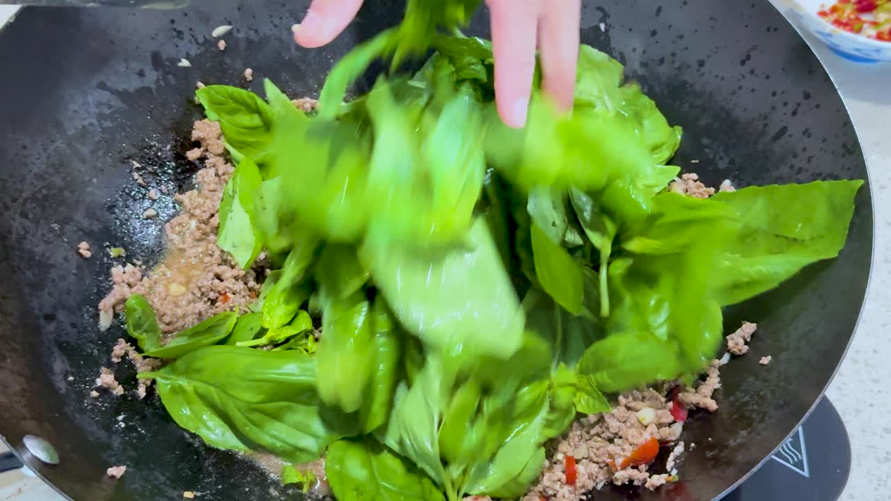 Fresh basil leaves are added to minced meat in a wok and stir-fried under bright kitchen lighting, with close-up overhead camera angles capturing the cooking process
