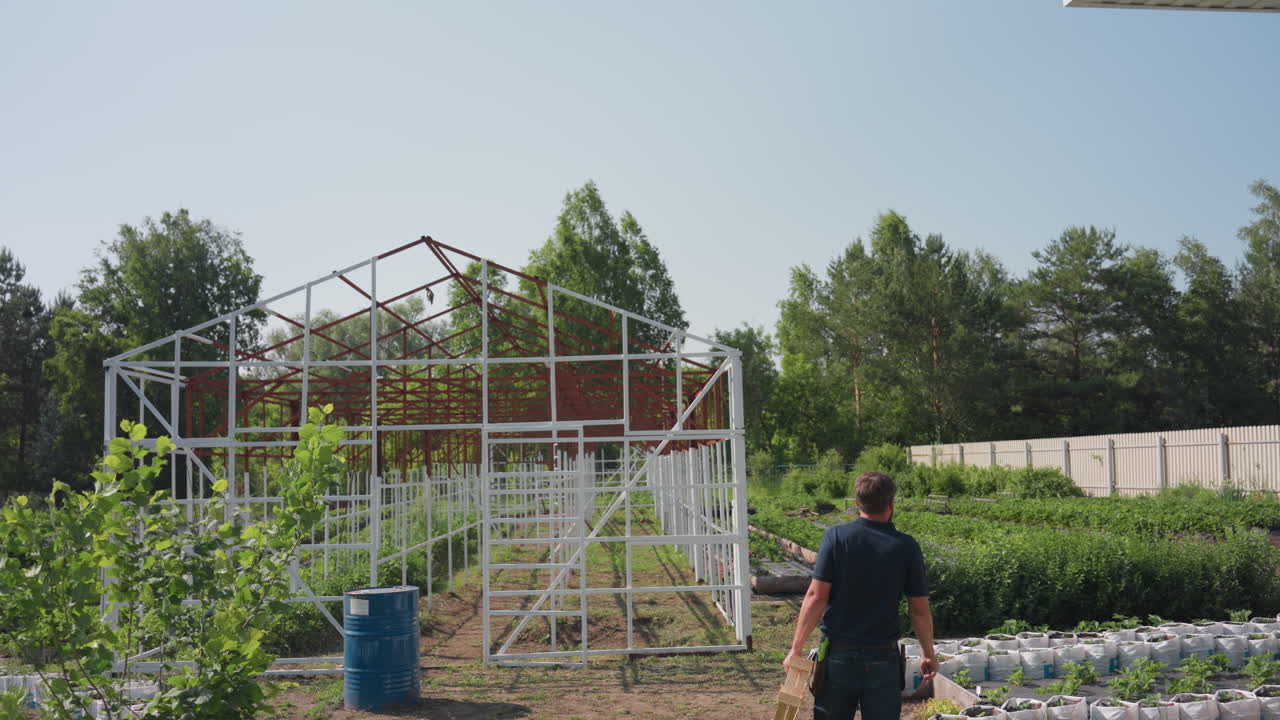 Rear view of farmer walking toward farm carrying wooden crate while inspecting greenhouse frame and plants on sunny day, surrounded by barrels grass trees and garden growth