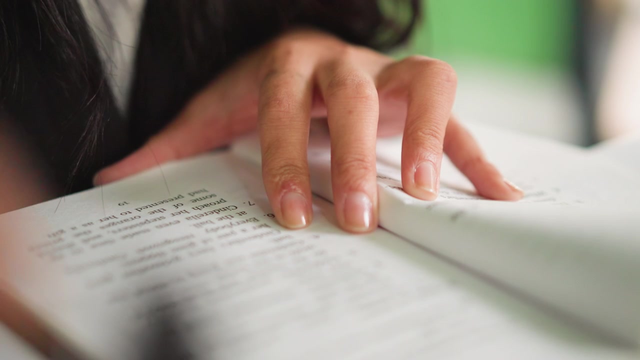 Primo piano delle mani di una giovane donna che riposano su un libro aperto con testo visibile e le dita che toccano delicatamente le pagine. La scena mostra una texture dettagliata e una luce soffusa a fuoco mentre lo sfondo rimane sfocato.