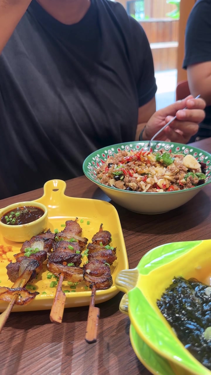 Seseorang berbaju hitam makan nasi dengan sayuran dan sate daging sapi panggang di restoran Bangkok yang terang benderang, menggunakan garpu dan sendok.