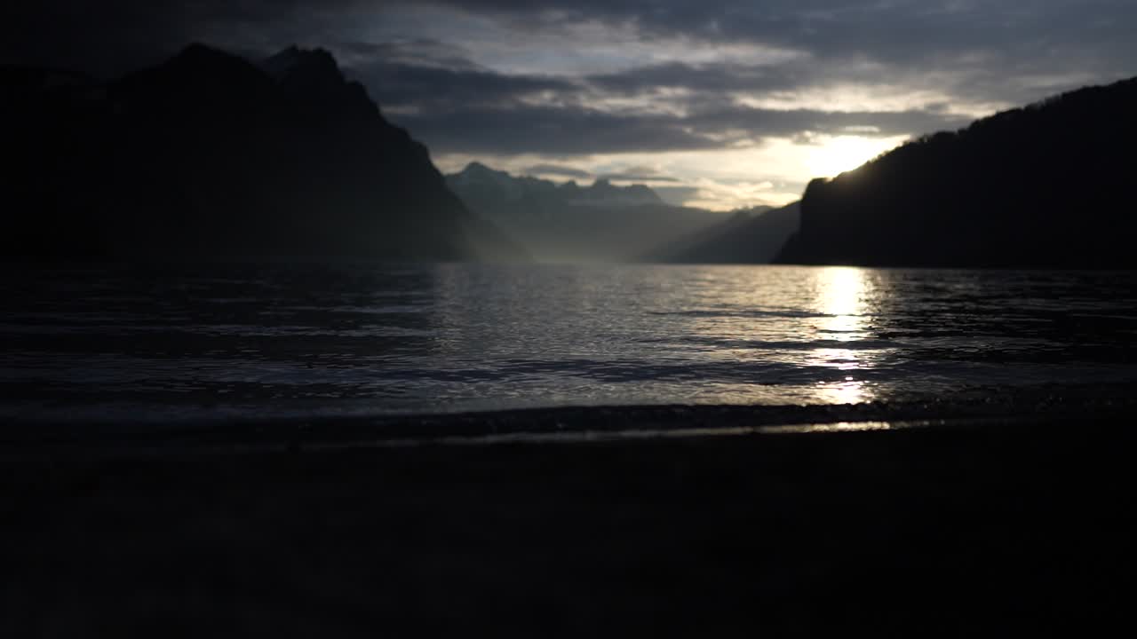 Gentle waves lap against the shore as the sun sets behind misty mountains over lake