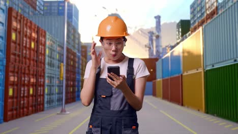 trabajadora asiática con gafas de protección y casco de seguridad mirando su teléfono inteligente, sonriendo, y diciendo wow mientras está de pie en el almacén del patio de contenedores