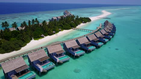 Traditional-bungalows-built-over-calm-shallow-lagoon-with-corals-and-pebbles-under-water,-close-to-tropical-beach-with-white-sand-in-Maldives