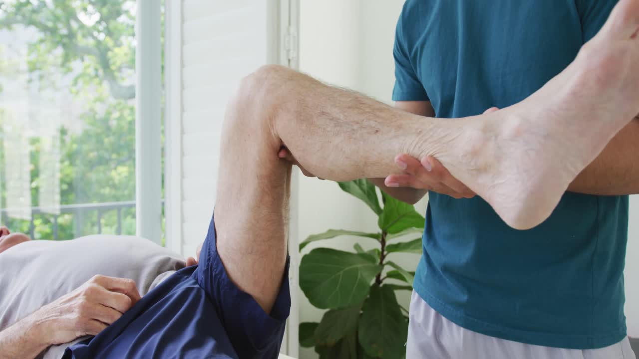 Biracial male therapist examining while lifting senior man's leg at health club