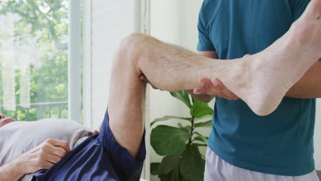 biracial male therapist examining while lifting senior man's leg at health club